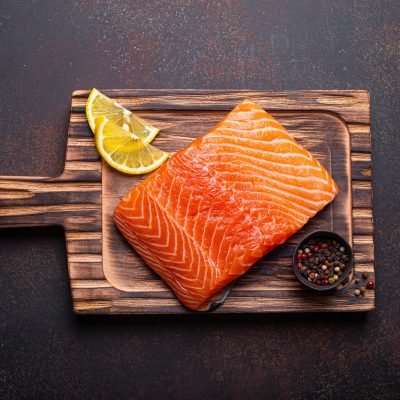 Fresh raw salmon fillet on wooden kitchen cutting board with seasonings and lemon top view on dark